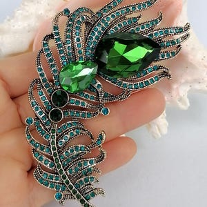 May include: A feather-shaped brooch crafted from antique silver-toned metal, featuring two large, faceted emerald-green teardrop crystals and numerous smaller, round teal-colored rhinestones embedded throughout the intricate, curved metal structure.
