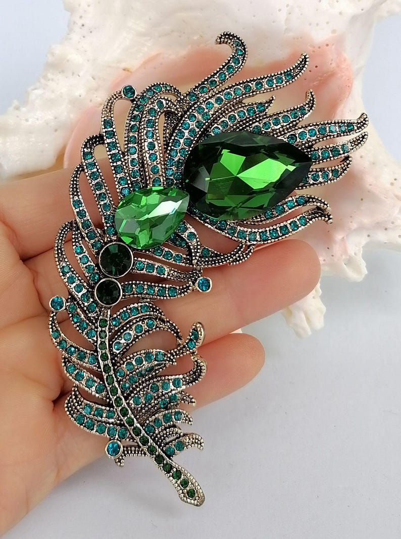 May include: A feather-shaped brooch crafted from antique silver-toned metal, featuring two large, faceted emerald-green teardrop crystals and numerous smaller, round teal-colored rhinestones embedded throughout the intricate, curved metal structure.