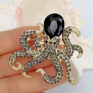 K&ouml;nnte beinhalten: Goldfarbene Oktopus-Brosche mit grauen Kristallakzenten und einem gro&szlig;en schwarzen Edelstein. Die Brosche hat detaillierte Tentakel und einen zentralen schwarzen Edelstein, was ein auff&auml;lliges Accessoire ergibt.