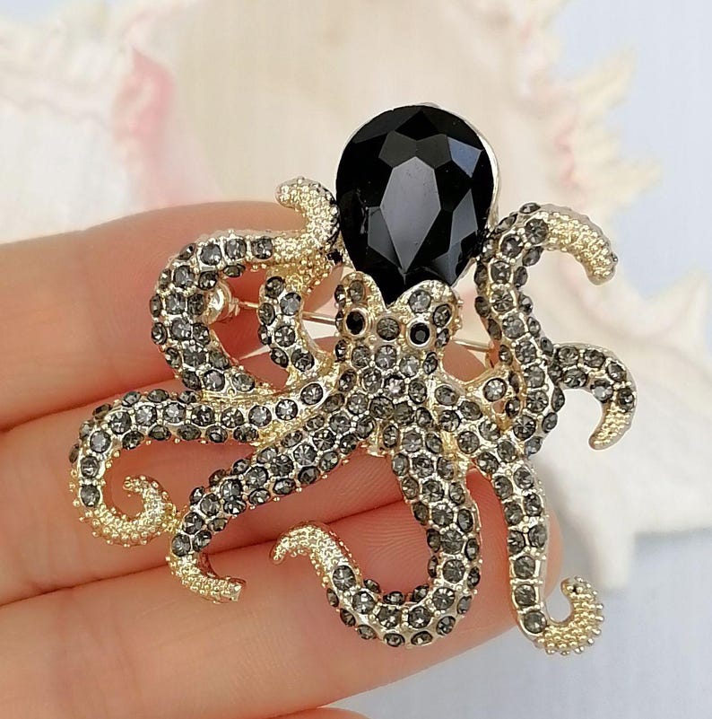 K&ouml;nnte beinhalten: Goldfarbene Oktopus-Brosche mit grauen Kristallakzenten und einem gro&szlig;en schwarzen Edelstein. Die Brosche hat detaillierte Tentakel und einen zentralen schwarzen Edelstein, was ein auff&auml;lliges Accessoire ergibt.