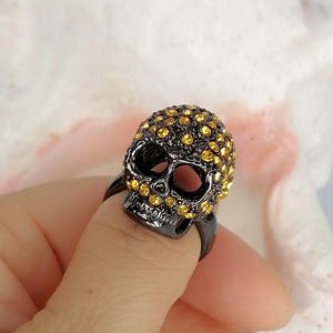 Op de afbeelding: Een zwarte metalen ring met een driedimensionaal schedelontwerp. Het oppervlak van de schedel is ingelegd met talloze kleine, ronde, goudgele kristallen die de schedel en oogkassen bedekken, terwijl de kaak glad en donker van kleur blijft.