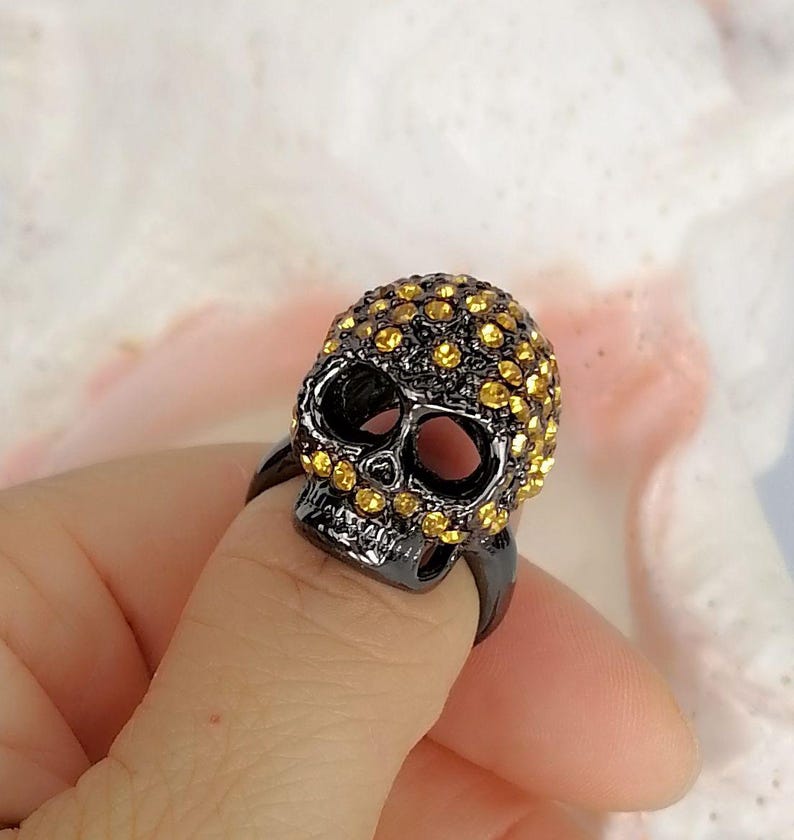 Op de afbeelding: Een zwarte metalen ring met een driedimensionaal schedelontwerp. Het oppervlak van de schedel is ingelegd met talloze kleine, ronde, goudgele kristallen die de schedel en oogkassen bedekken, terwijl de kaak glad en donker van kleur blijft.
