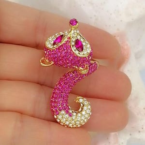K&ouml;nnte beinhalten: Eine fuchsformige Brosche aus goldfarbenem Metall, die vollst&auml;ndig mit leuchtend rosa und klaren wei&szlig;en Kristallen besetzt ist. Das Design zeigt zwei marquise-geschliffene fuchsiafarbene Steine als Augen und einen runden Stein am Kopf.