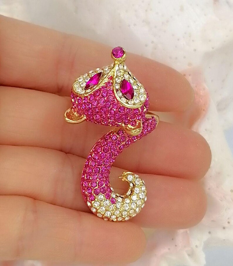 K&ouml;nnte beinhalten: Eine fuchsformige Brosche aus goldfarbenem Metall, die vollst&auml;ndig mit leuchtend rosa und klaren wei&szlig;en Kristallen besetzt ist. Das Design zeigt zwei marquise-geschliffene fuchsiafarbene Steine als Augen und einen runden Stein am Kopf.