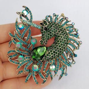 May include: A gold-toned brooch in the shape of a unicorn. The unicorn's body is covered in green rhinestones, with teal and iridescent accents. A large green gemstone is at the center. The brooch is held in a hand.
