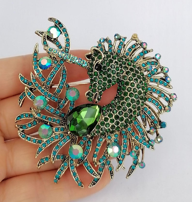 May include: A gold-toned brooch in the shape of a unicorn. The unicorn's body is covered in green rhinestones, with teal and iridescent accents. A large green gemstone is at the center. The brooch is held in a hand.