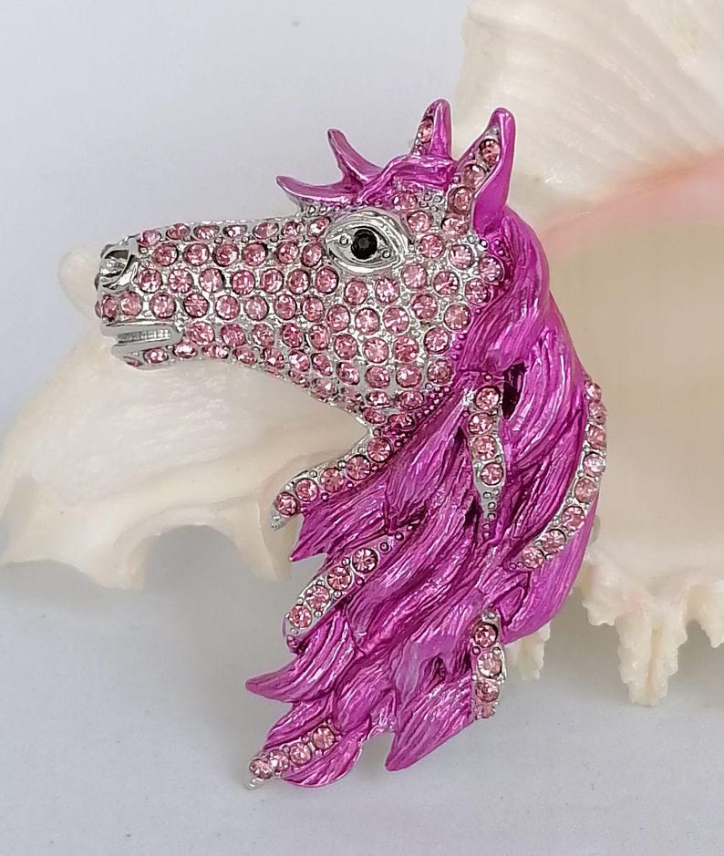 K&ouml;nnte beinhalten: Eine pinkfarbene und silberfarbene Brosche in Form eines Pferdekopfes. Der Pferdekopf ist mit rosa Strasssteinen besetzt, mit einer silberfarbenen Metallnase und einem Auge. Die M&auml;hne ist leuchtend pink mit Strassakzenten.