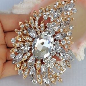 May include: A gold-tone brooch featuring a large central oval clear crystal surrounded by smaller marquise and round clear crystals arranged in a leaf-like pattern. The metal setting is intricate and filigree-style, holding the faceted stones.