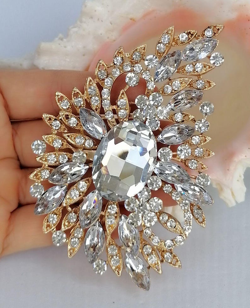 May include: A gold-tone brooch featuring a large central oval clear crystal surrounded by smaller marquise and round clear crystals arranged in a leaf-like pattern. The metal setting is intricate and filigree-style, holding the faceted stones.