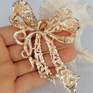May include: A gold-colored bow brooch with intricate filigree detailing. The brooch has a pin on the back for secure attachment. The bow design features a layered appearance with delicate cut-out patterns, creating a textured and elegant accessory.
