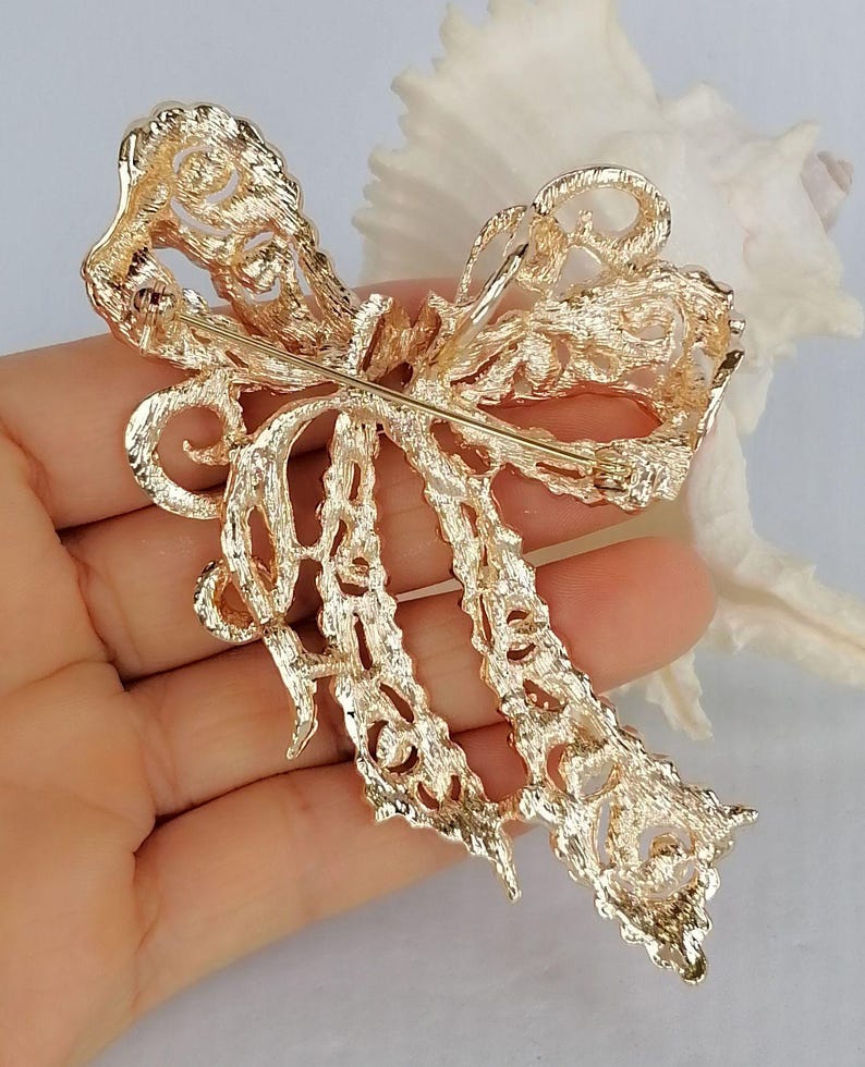 May include: A gold-colored bow brooch with intricate filigree detailing. The brooch has a pin on the back for secure attachment. The bow design features a layered appearance with delicate cut-out patterns, creating a textured and elegant accessory.