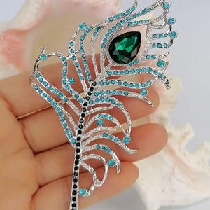 May include: A silver peacock feather brooch with teal and black detailing. The brooch has teal gemstones and a large, emerald-green teardrop-shaped stone. The feather design is intricate and elegant.