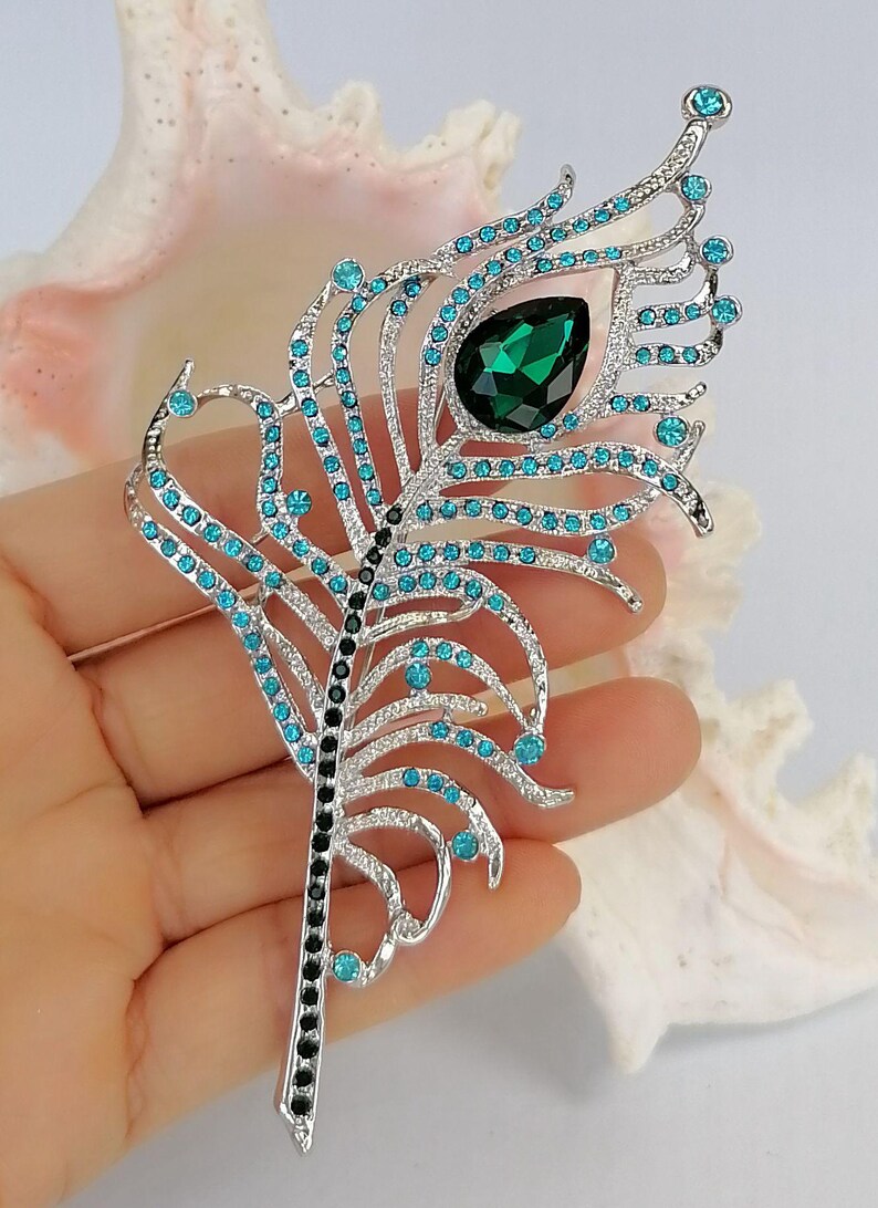 May include: A silver peacock feather brooch with teal and black detailing. The brooch has teal gemstones and a large, emerald-green teardrop-shaped stone. The feather design is intricate and elegant.