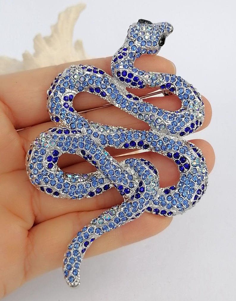 May include: A silver snake brooch, adorned with blue rhinestones in various shades. The snake's head is black, and the body is coiled. This decorative brooch is a striking piece of jewellery.