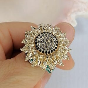 K&ouml;nnte beinhalten: Ein sonnenblumenf&ouml;rmiger Ring mit goldfarbenen Bl&uuml;tenbl&auml;ttern in strukturierter Optik. Die Mitte der Blume ist dicht mit kleinen, dunkelgrauen, facettierten Steinen besetzt. Kleine, leuchtend gr&uuml;ne Steine sind am Ansatz der goldenen Bl&uuml;tenbl&auml;tter sichtbar.