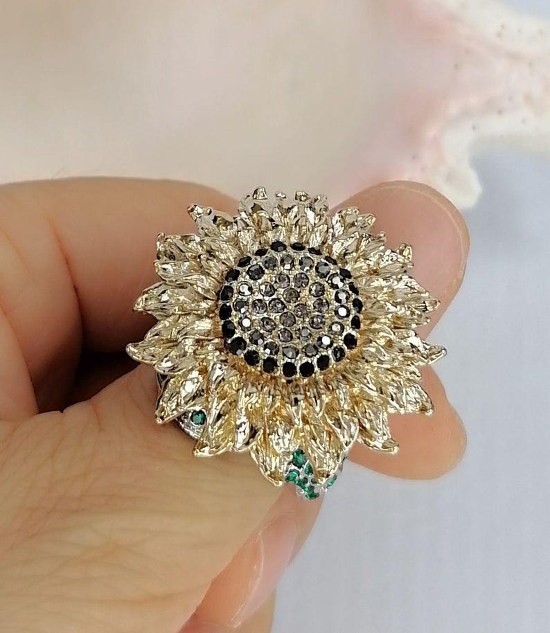 K&ouml;nnte beinhalten: Ein sonnenblumenf&ouml;rmiger Ring mit goldfarbenen Bl&uuml;tenbl&auml;ttern in strukturierter Optik. Die Mitte der Blume ist dicht mit kleinen, dunkelgrauen, facettierten Steinen besetzt. Kleine, leuchtend gr&uuml;ne Steine sind am Ansatz der goldenen Bl&uuml;tenbl&auml;tter sichtbar.