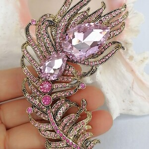 May include: A feather-shaped brooch crafted from antique gold-toned metal, featuring two large, faceted, pear-shaped light pink crystals and numerous smaller, round, vibrant pink rhinestones embedded throughout the intricate, curved metal structure.