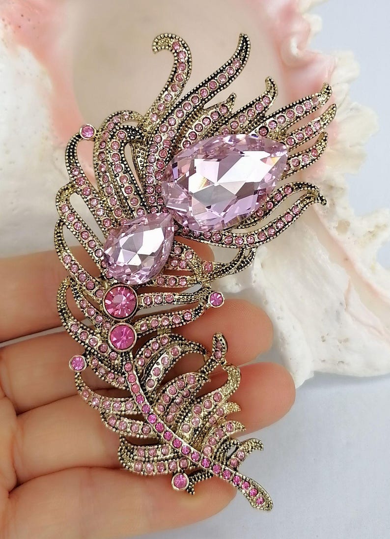 May include: A feather-shaped brooch crafted from antique gold-toned metal, featuring two large, faceted, pear-shaped light pink crystals and numerous smaller, round, vibrant pink rhinestones embedded throughout the intricate, curved metal structure.