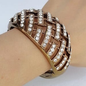 Puede incluir: Una pulsera brazalete ancha de metal en tono bronce con un diseño geométrico de celosía calada. La superficie de las barras metálicas está incrustada con numerosas piedras de estrás pequeñas, transparentes y de corte redondo.