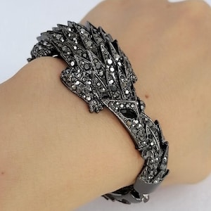 May include: A dark gunmetal-toned bracelet shaped like a stylized lizard or reptile. The entire surface of the creature is encrusted with numerous small, sparkling black crystals, creating a textured, reflective appearance across the body.