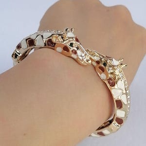 May include: A gold-tone hinged bangle bracelet featuring two giraffe heads meeting at the center. The bracelet is decorated with white and brown enamel patches mimicking a giraffe's coat pattern and is accented with small clear crystals.