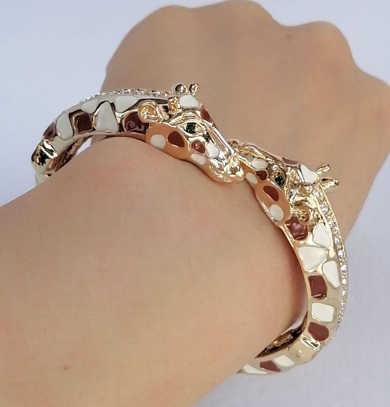 May include: A gold-tone hinged bangle bracelet featuring two giraffe heads meeting at the center. The bracelet is decorated with white and brown enamel patches mimicking a giraffe's coat pattern and is accented with small clear crystals.