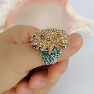 K&ouml;nnte beinhalten: Ein silberfarbener Ring mit einem gro&szlig;en Sonnenblumenmotiv. Die Mitte der Blume ist mit kleinen bernsteinfarbenen Steinen besetzt, w&auml;hrend die Bl&uuml;tenbl&auml;tter aus strukturiertem goldfarbenem Metall bestehen. Der Ring ist mit kleinen gr&uuml;nen Steinen verziert.