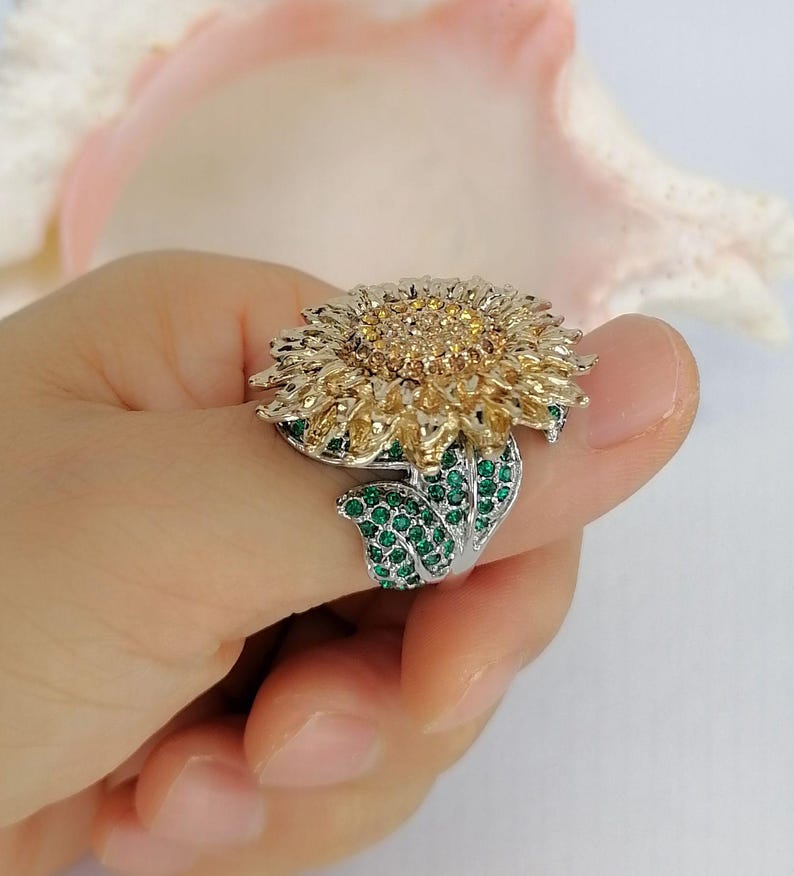 K&ouml;nnte beinhalten: Ein silberfarbener Ring mit einem gro&szlig;en Sonnenblumenmotiv. Die Mitte der Blume ist mit kleinen bernsteinfarbenen Steinen besetzt, w&auml;hrend die Bl&uuml;tenbl&auml;tter aus strukturiertem goldfarbenem Metall bestehen. Der Ring ist mit kleinen gr&uuml;nen Steinen verziert.