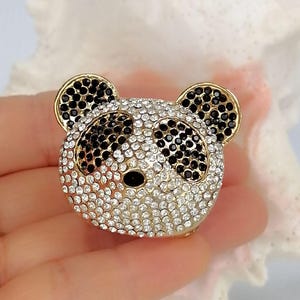 May include: A panda bear head brooch featuring a gold-tone metal frame encrusted with clear white rhinestones across the face and ears, with black rhinestones forming the eye patches, ears, and nose, creating a detailed animal-shaped accessory.