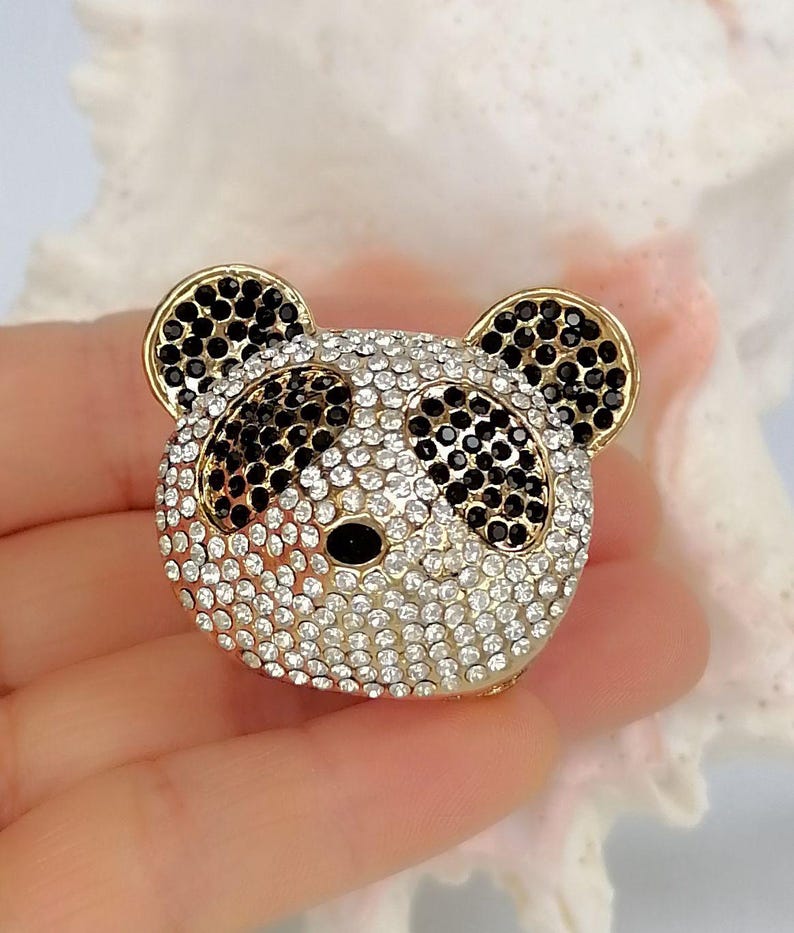 May include: A panda bear head brooch featuring a gold-tone metal frame encrusted with clear white rhinestones across the face and ears, with black rhinestones forming the eye patches, ears, and nose, creating a detailed animal-shaped accessory.