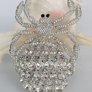 May include: A silver-coloured brooch in the shape of a crab, adorned with many clear crystals. The crab features black eyes and articulated legs. The brooch is set against a white background.