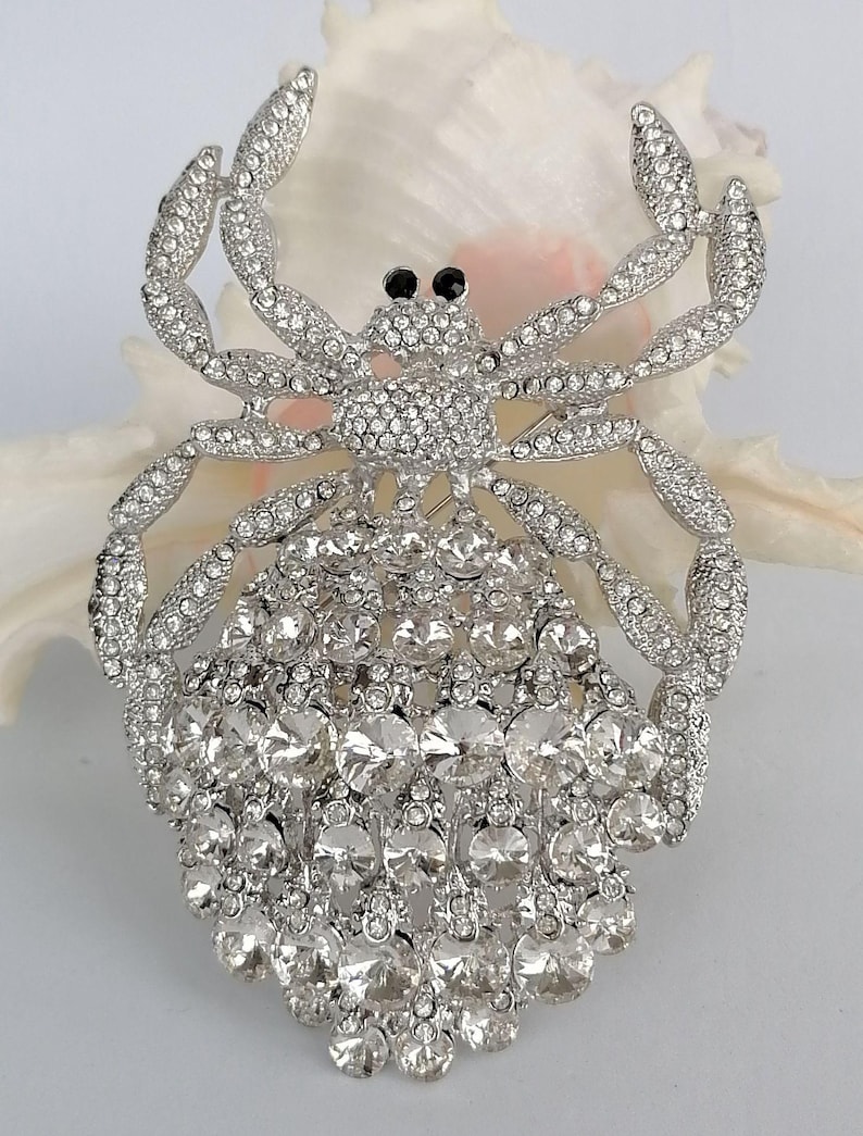 May include: A silver-coloured brooch in the shape of a crab, adorned with many clear crystals. The crab features black eyes and articulated legs. The brooch is set against a white background.