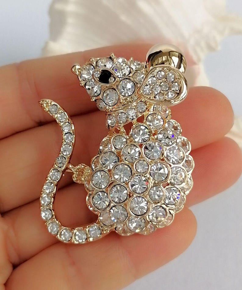 May include: A gold-toned mouse brooch encrusted with clear, sparkling crystals. The mouse has a rounded body, a long, curved tail, and a small, black eye. The brooch is designed to be worn as a decorative accessory.