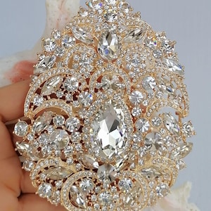 May include: A gold-toned, ornate brooch encrusted with numerous clear, sparkling crystals. The brooch features a large, central teardrop-shaped crystal surrounded by smaller, various-sized crystals. The design is symmetrical and detailed, with a vintage or bridal aesthetic.