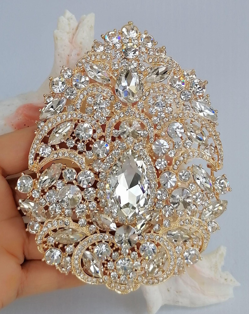 May include: A gold-toned, ornate brooch encrusted with numerous clear, sparkling crystals. The brooch features a large, central teardrop-shaped crystal surrounded by smaller, various-sized crystals. The design is symmetrical and detailed, with a vintage or bridal aesthetic.