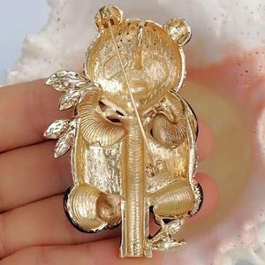 May include: A gold-toned metal brooch shaped like a panda bear holding a bamboo stalk. The reverse side features a textured finish with a horizontal pin mechanism and a secure clasp. The design shows the panda's head, limbs, and body in relief.