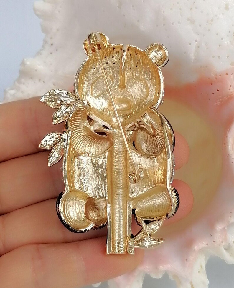 May include: A gold-toned metal brooch shaped like a panda bear holding a bamboo stalk. The reverse side features a textured finish with a horizontal pin mechanism and a secure clasp. The design shows the panda's head, limbs, and body in relief.