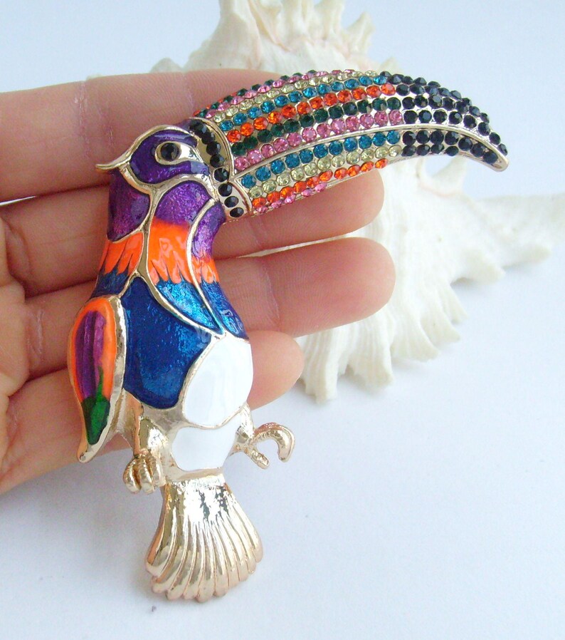 May include: A colorful toucan brooch with a gold-tone finish. The bird features a vibrant design with a rainbow-colored beak, purple head, orange chest, and blue body. The brooch is an eye-catching accessory.