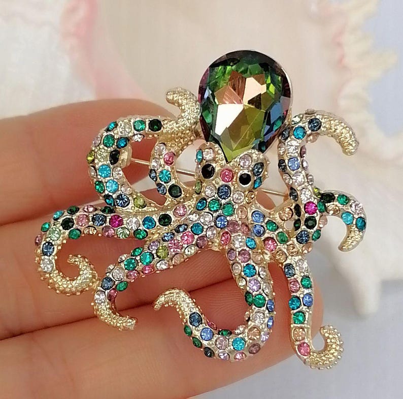 K&ouml;nnte beinhalten: Goldfarbene Oktopus-Brosche mit bunten Strasssteinen besetzt. Die Brosche hat einen gro&szlig;en, birnenf&ouml;rmigen gr&uuml;nen Kristall oben und ist mit verschiedenfarbigen Strasssteinen verziert, darunter Blau, Rosa und Gr&uuml;n. Die Tentakel des Oktopus sind gekr&auml;uselt.
