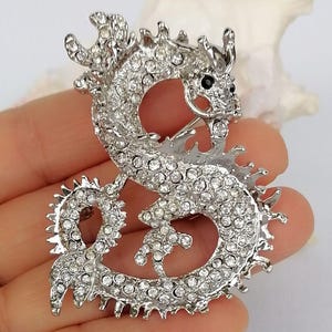 Puede incluir: Broche de drag&oacute;n plateado, adornado con brillantes transparentes. El drag&oacute;n est&aacute; representado en una postura curvada, con la cabeza girada y ojos negros. Este broche es una joya, perfecto para a&ntilde;adir un toque de fantas&iacute;a a un atuendo.