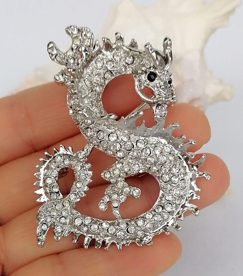 Puede incluir: Broche de drag&oacute;n plateado, adornado con brillantes transparentes. El drag&oacute;n est&aacute; representado en una postura curvada, con la cabeza girada y ojos negros. Este broche es una joya, perfecto para a&ntilde;adir un toque de fantas&iacute;a a un atuendo.