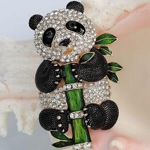May include: A panda brooch featuring clear rhinestones on the head and body, black textured enamel limbs and ears, and a green enamel bamboo stalk with leaves. The panda is holding the bamboo stalk, which is accented with small clear rhinestones.
