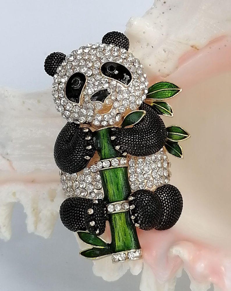 May include: A panda brooch featuring clear rhinestones on the head and body, black textured enamel limbs and ears, and a green enamel bamboo stalk with leaves. The panda is holding the bamboo stalk, which is accented with small clear rhinestones.