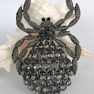 May include: A decorative spider brooch, crafted from dark grey metal and adorned with clear and grey crystals. The brooch features a detailed design with multiple crystals, creating a sparkling effect. The spider's body is a mix of round and teardrop-shaped crystals.