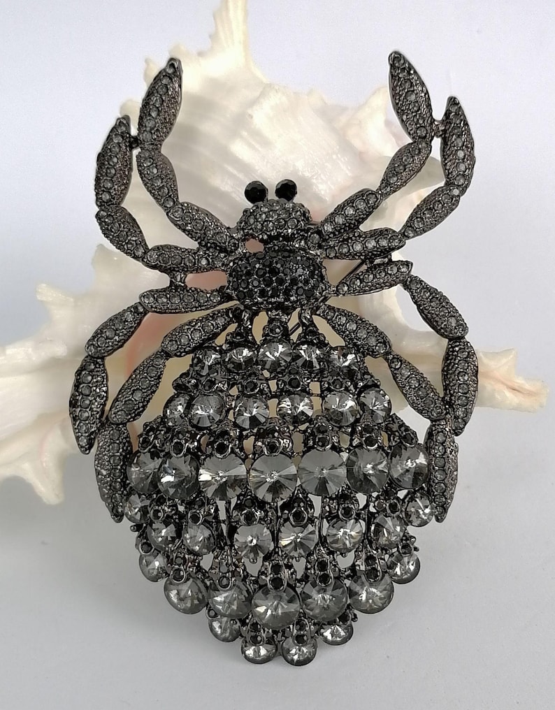 May include: A decorative spider brooch, crafted from dark grey metal and adorned with clear and grey crystals. The brooch features a detailed design with multiple crystals, creating a sparkling effect. The spider's body is a mix of round and teardrop-shaped crystals.