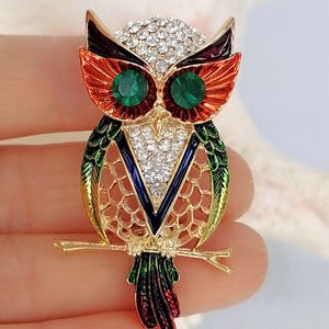 Op de afbeelding: Een uilenbroche van goudkleurig metaal met levendige oranje, groene en blauwe emaille details. De kop en borst van de uil zijn versierd met heldere, fonkelende strass-steentjes, terwijl de ogen zijn ingezet met twee ronde groene kristallen.