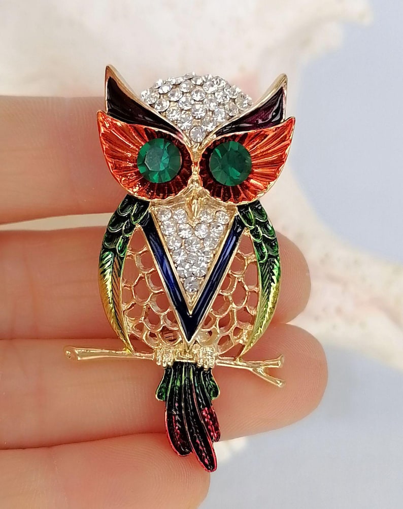Op de afbeelding: Een uilenbroche van goudkleurig metaal met levendige oranje, groene en blauwe emaille details. De kop en borst van de uil zijn versierd met heldere, fonkelende strass-steentjes, terwijl de ogen zijn ingezet met twee ronde groene kristallen.