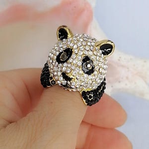 Peut inclure: Une bague en forme de t&ecirc;te de panda, dot&eacute;e d'une base en m&eacute;tal dor&eacute; recouverte de strass blancs transparents. Le panda pr&eacute;sente des accents en &eacute;mail noir autour des yeux, des oreilles et des pattes, avec de petits cristaux noirs incrust&eacute;s.
