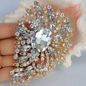 May include: A gold-toned brooch featuring a large central oval clear crystal surrounded by smaller marquise and round clear crystals arranged in a leaf-like pattern. The metal setting is intricate and filigree-inspired, showcasing a symmetrical design.
