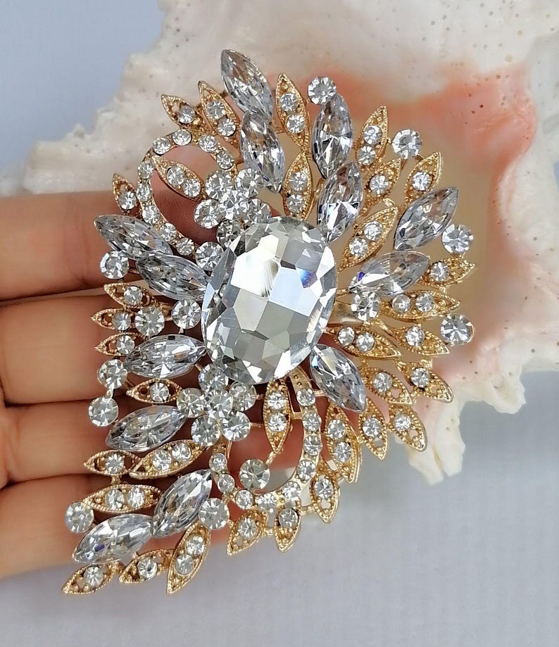 May include: A gold-toned brooch featuring a large central oval clear crystal surrounded by smaller marquise and round clear crystals arranged in a leaf-like pattern. The metal setting is intricate and filigree-inspired, showcasing a symmetrical design.
